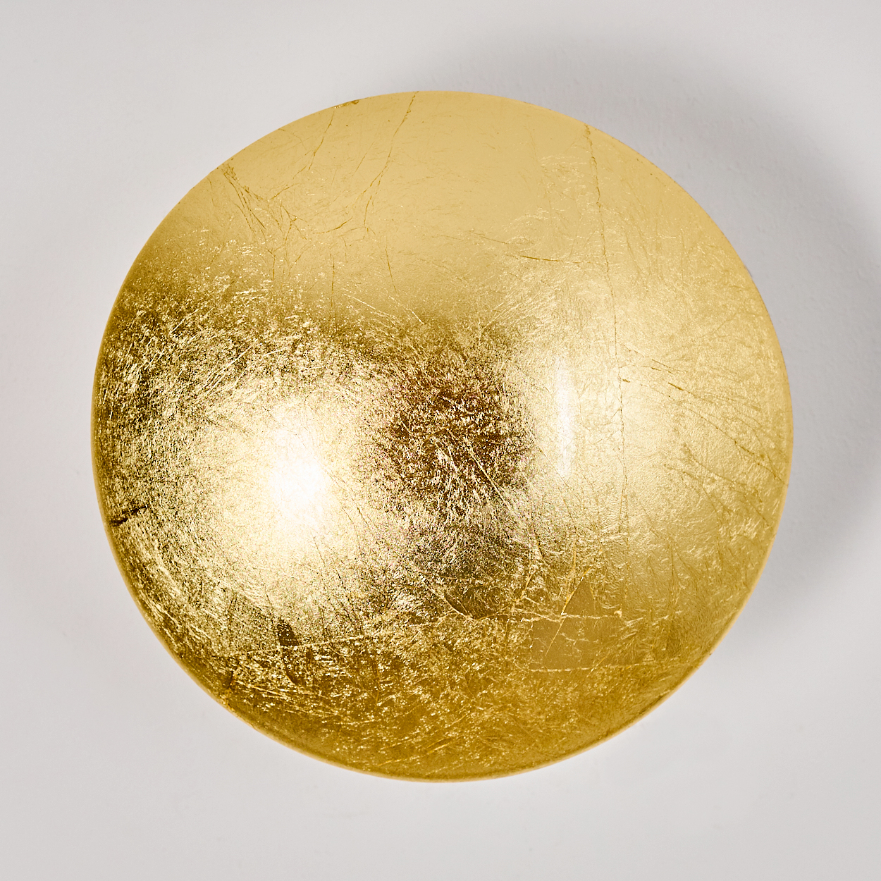Ein rundes, goldenes Objekt mit strukturierter Oberfläche, möglicherweise aus Blattgold, vor weißem Hintergrund.