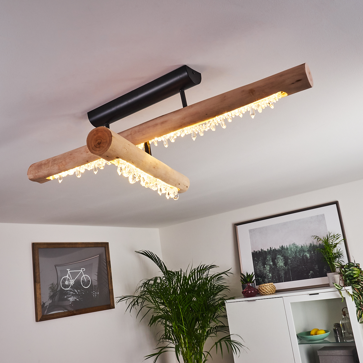 Eine moderne Deckenleuchte mit Holzbalken und Kristallakzenten. Kunstwerke und Pflanzen sind im Hintergrund zu sehen, zusammen mit einem weißen Schrank.