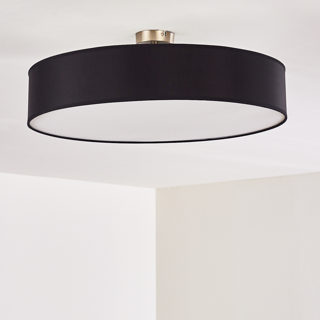 Eine kreisförmige Deckenlampe. Sie hat einen schwarzen Schirm und ein weißes Innenleben mit Metallbasis. Der Hintergrund zeigt weiße Wände.