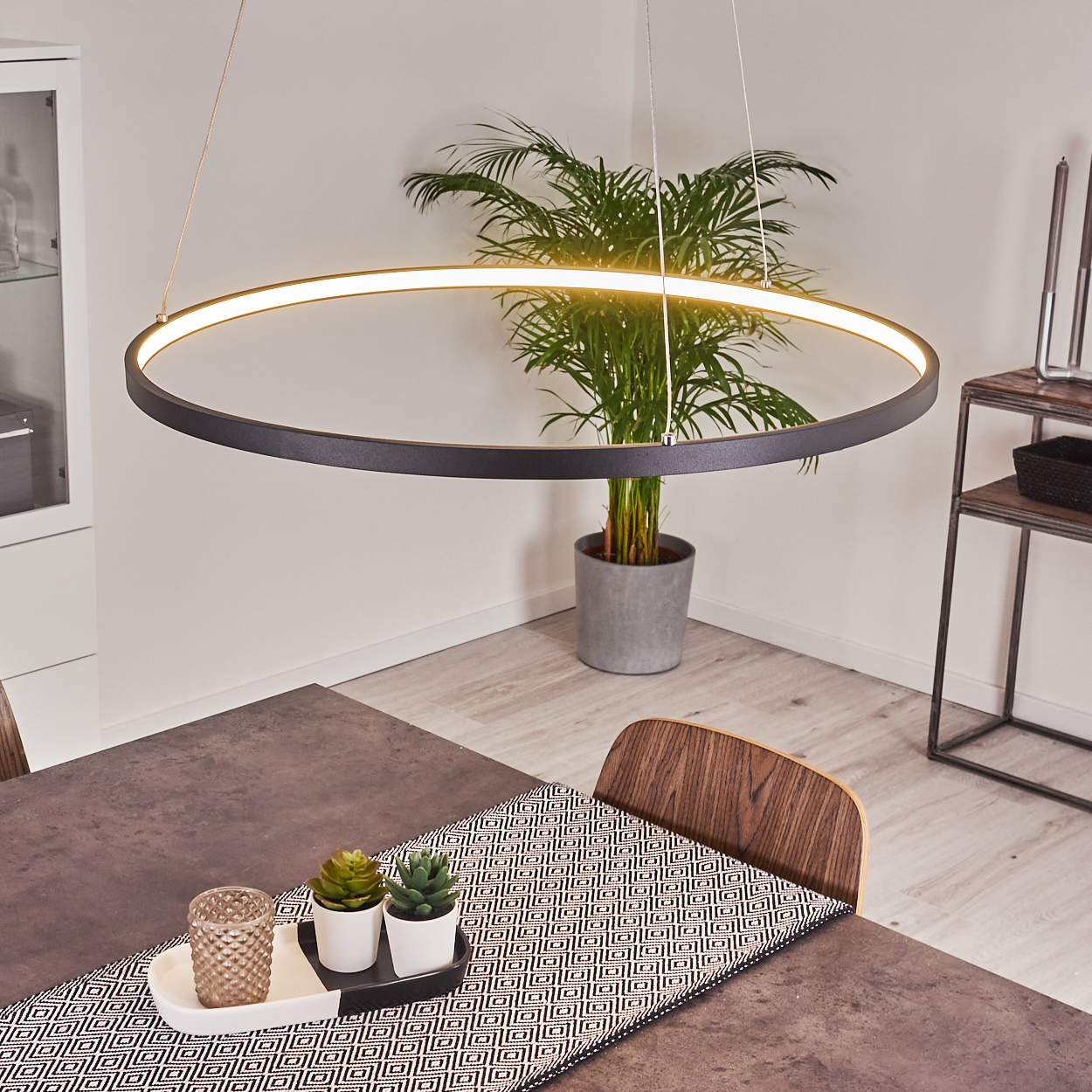 Ein modernes Esszimmer mit einer kreisförmigen Leuchte, einer Pflanze und einem Tisch mit dekorativen Gegenständen. Licht und Pflanze im Hintergrund.