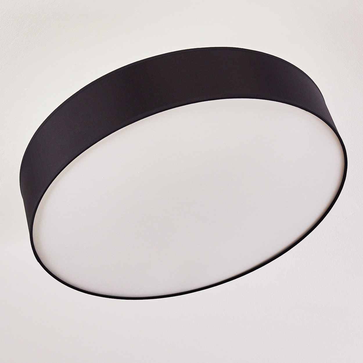 Eine schwarze, kreisförmige Deckenlampe mit weißem Innenraum ist an einer weißen Decke angebracht. Das Design der Lampe ist modern und schlicht.