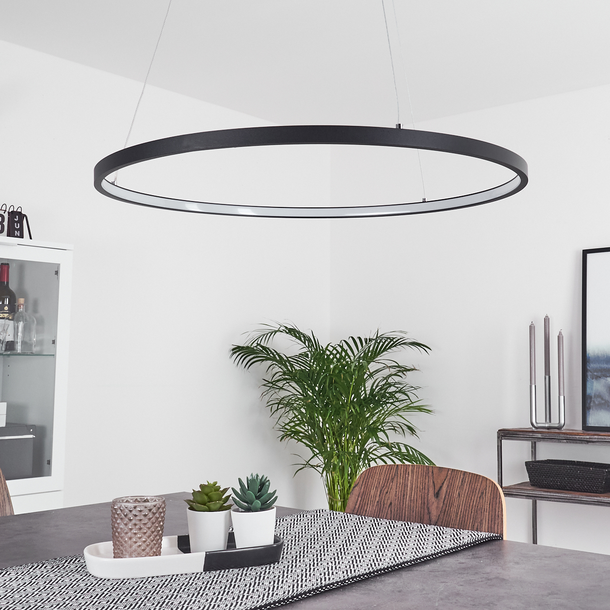 Ein modernes Esszimmer mit einer großen schwarzen kreisförmigen Lampe, einem Tisch mit Dekoration, einer Pflanze und einem Holzstuhl.