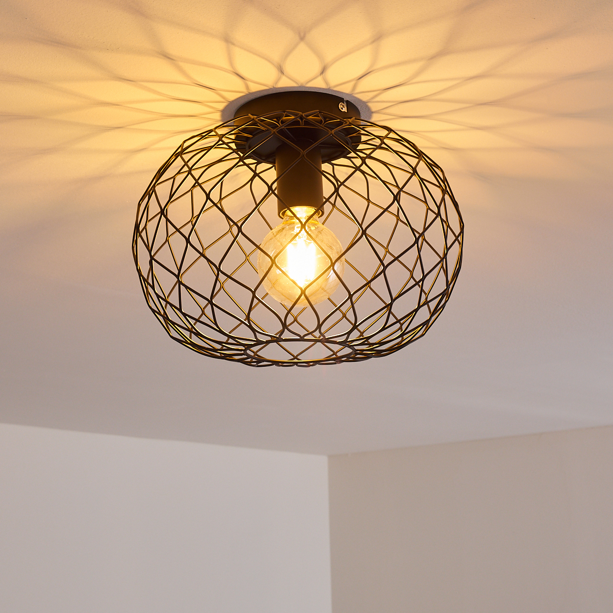 Eine schwarze Drahtlampe hängt von der Decke. Die Glühbirne leuchtet und wirft ein Muster auf die Decke. Im Hintergrund sind weiße Wände.