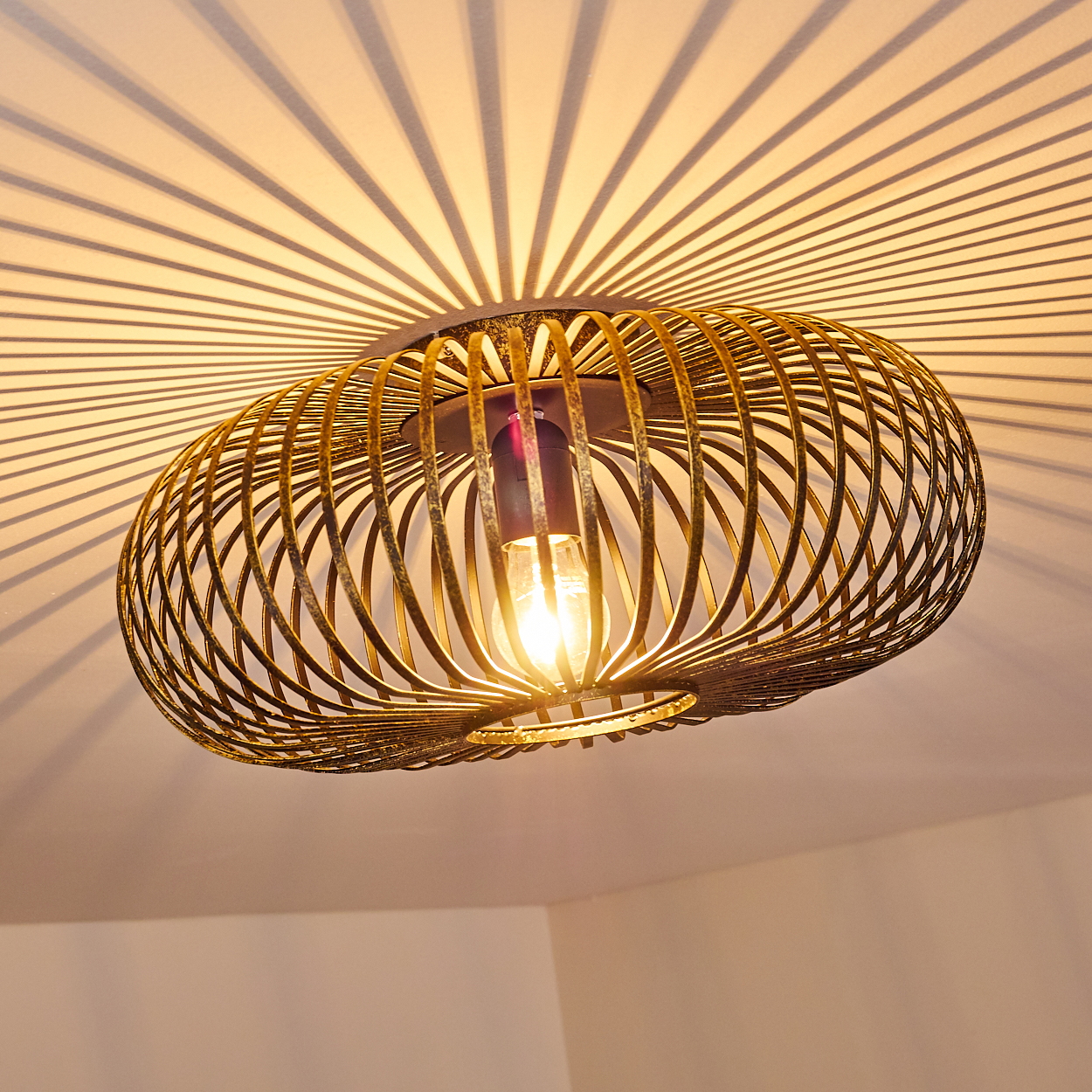 Eine goldene, käfigartige Deckenlampe mit eingeschalteter Glühbirne wirft radiale Schatten auf die Decke. Die Zimmerecke ist sichtbar.
