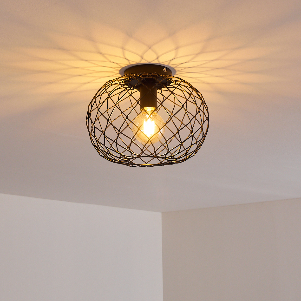Eine schwarze Deckenlampe mit Drahtrahmen und einer leuchtenden Glühbirne, die einen gemusterten Schatten auf die weiße Decke wirft. Weiße Wände in den Ecken.