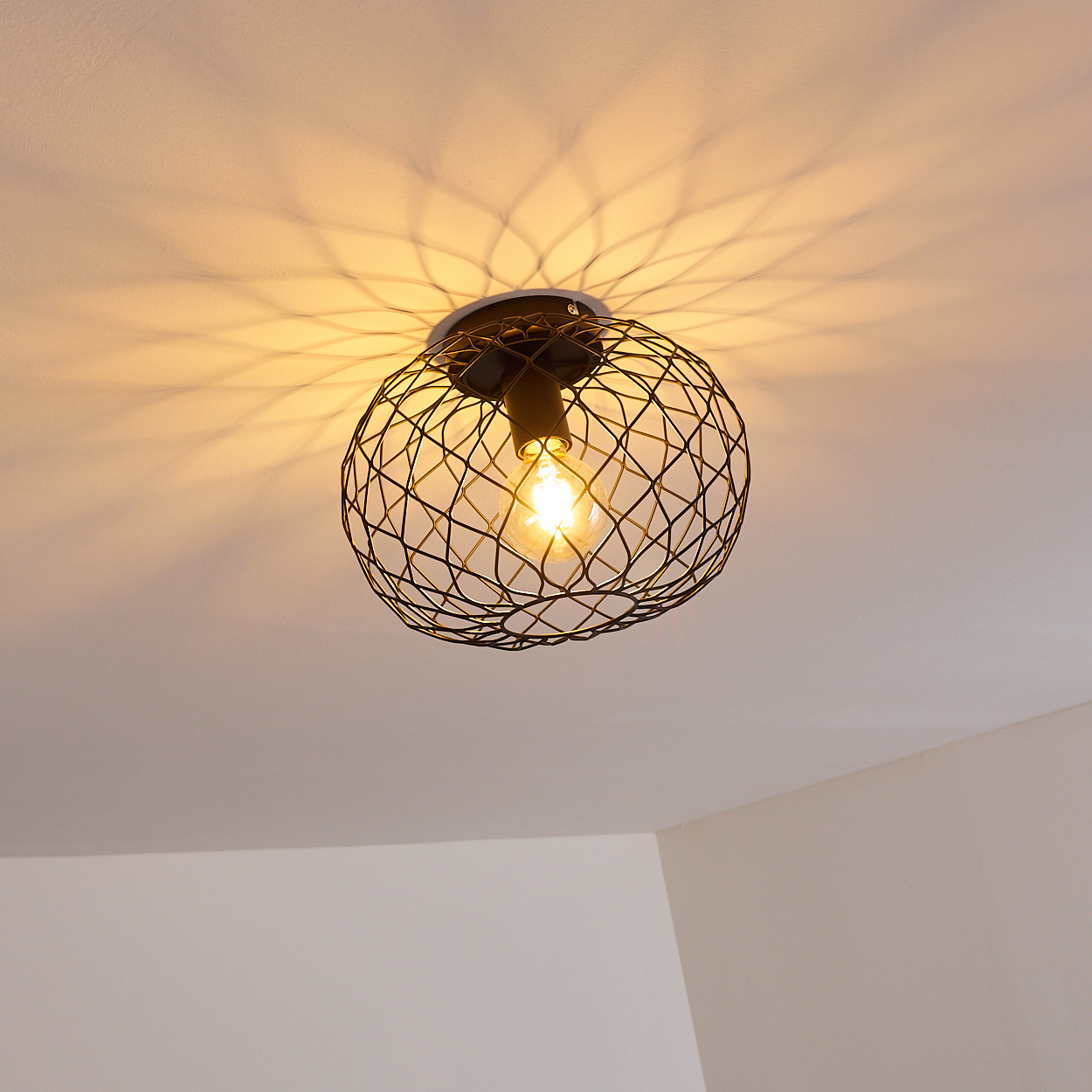 Eine schwarze Metallkäfig-Leuchte mit einer leuchtenden Glühbirne hängt von der Decke. Die Decke ist weiß, das Licht wirft ein Muster darauf.