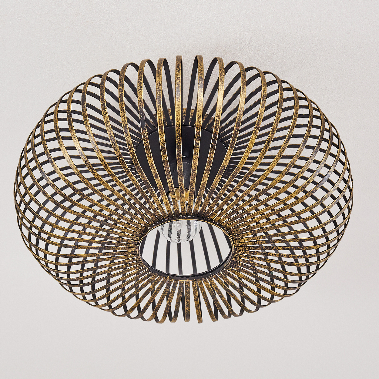 Nahaufnahme einer Deckenlampe. Es ist ein Metallkäfig mit goldenen und schwarzen Ringen und einer Glühbirne in der Mitte. Der Hintergrund ist eine weiße Decke.