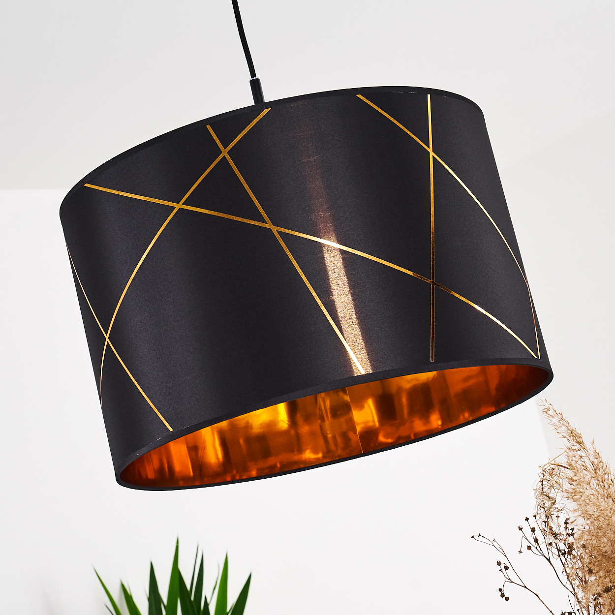Ein schwarzer Lampenschirm mit goldenen geometrischen Linien hängt. Das Innere ist goldfarben. Pflanzen sind im Hintergrund sichtbar.