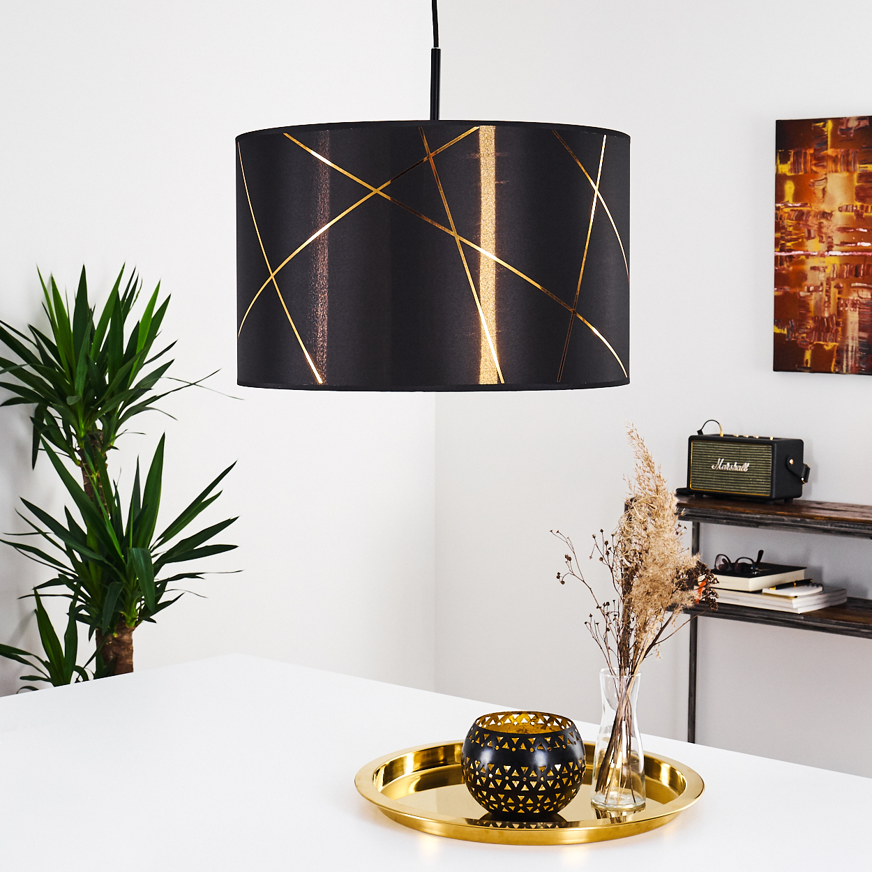 Ein schwarzer Lampenschirm mit goldenen Linien hängt über einer goldenen Schale mit einem Kerzenhalter und einer Vase. Ein Marshall-Lautsprecher steht auf einem Regal.