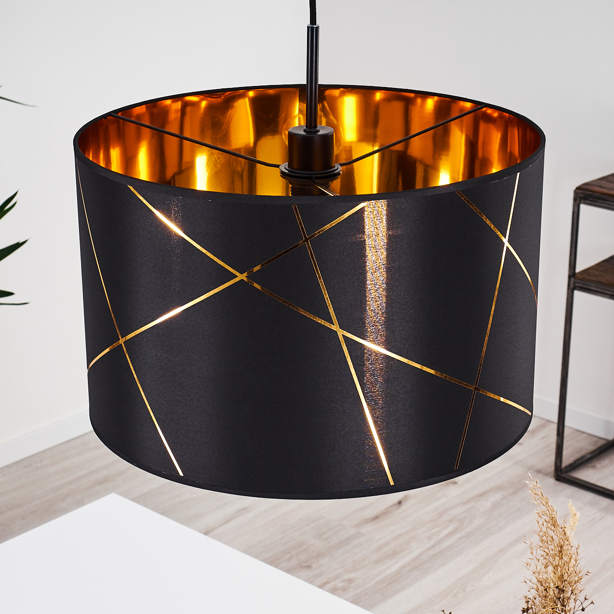 Ein schwarzer Lampenschirm mit goldenen Akzenten hängt von der Decke. Die Innenseite ist goldfarben. Ein Tisch und eine Pflanze sind im Hintergrund.