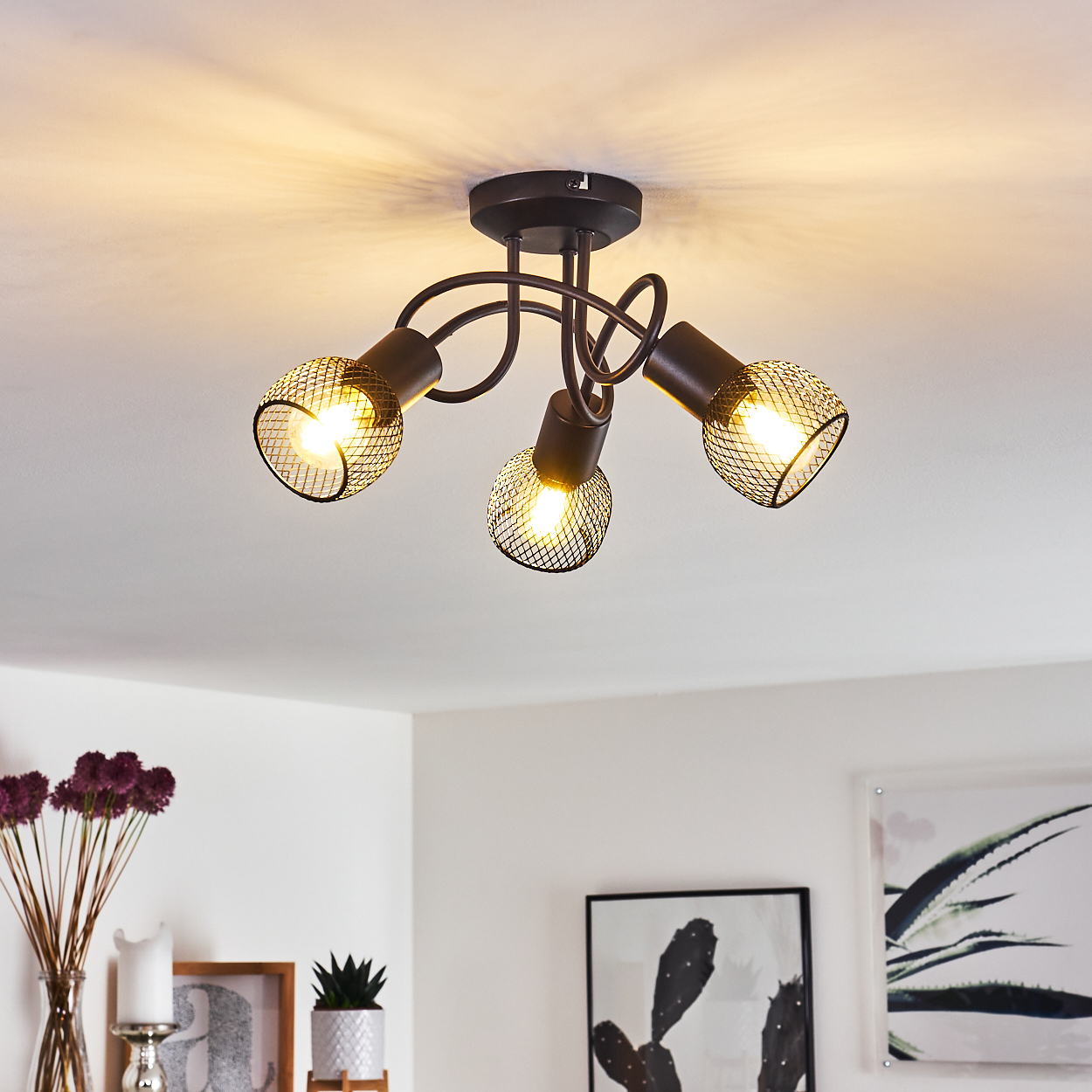 Deckenleuchte mit drei Lichtern, schwarzem Metallrahmen und Netzabdeckungen. Die Lichter sind an und werfen Schatten auf die weiße Decke.