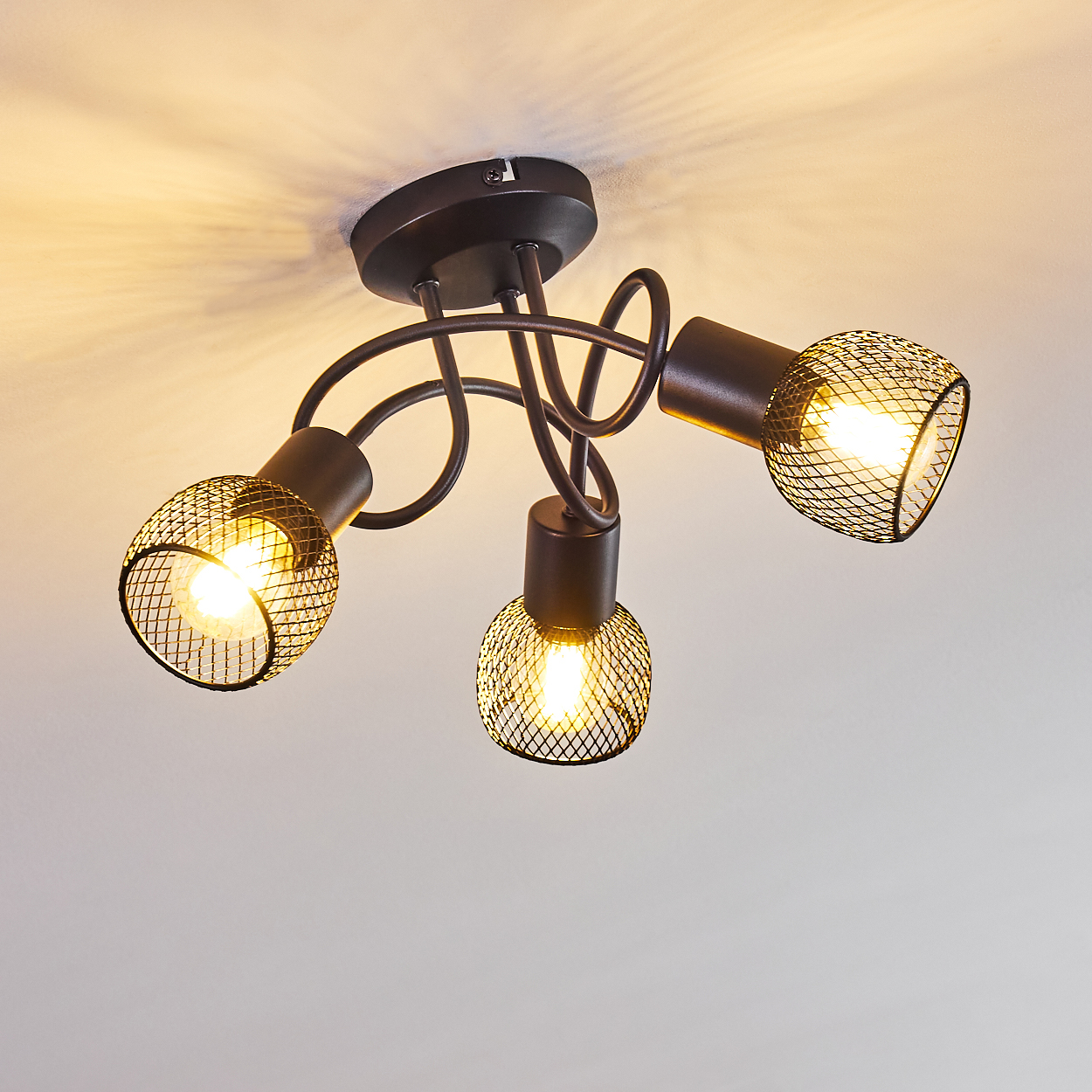 Eine Deckenleuchte mit drei Lampen. Jede Lampe ist von einem Drahtgeflecht umgeben. Die Leuchte ist dunkelbraun vor einer weißen Decke.