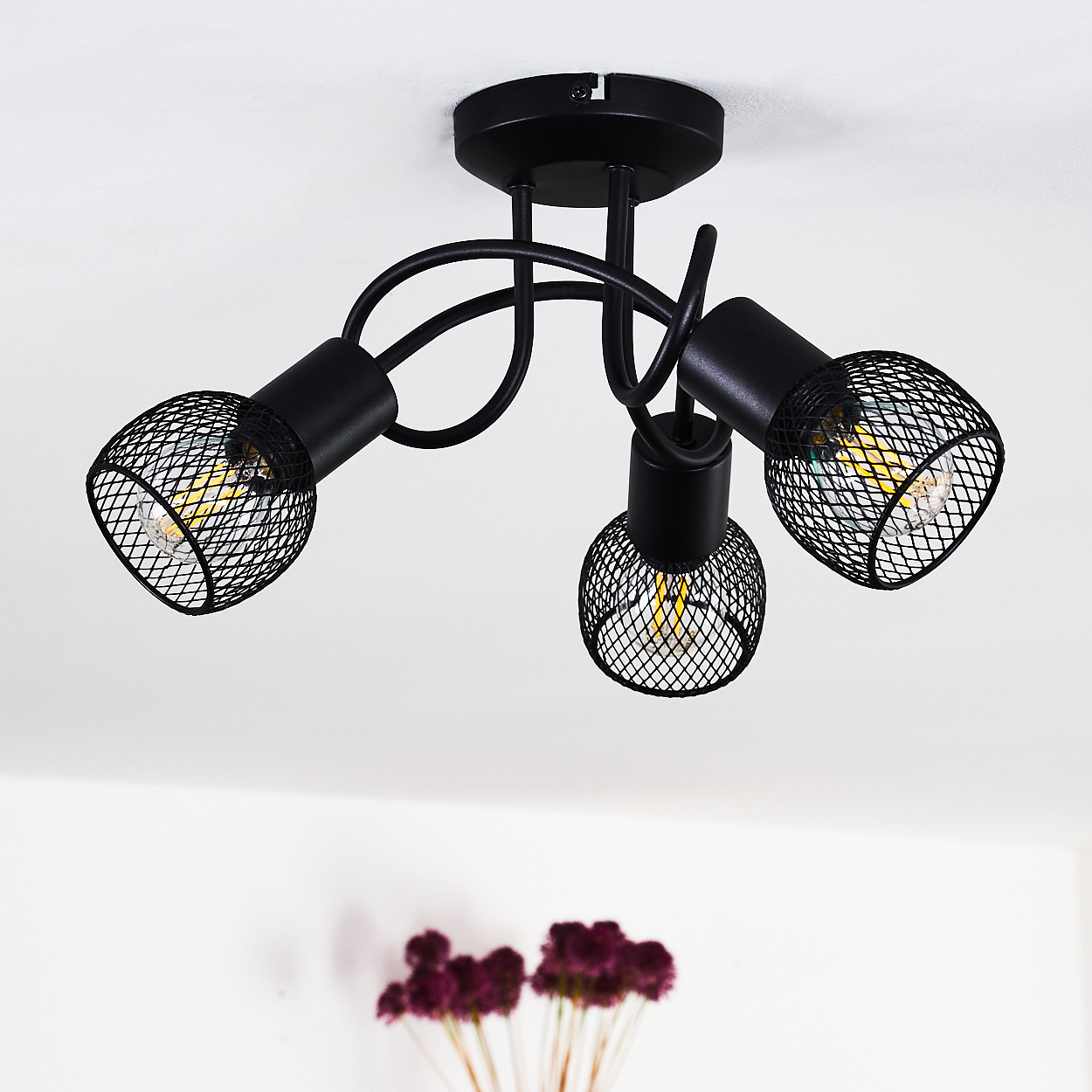 Eine schwarze Deckenleuchte mit drei Lampen mit Netzabdeckung. Glühbirnen sind sichtbar. Der Hintergrund ist weiß, mit einem Hauch von Blumen.