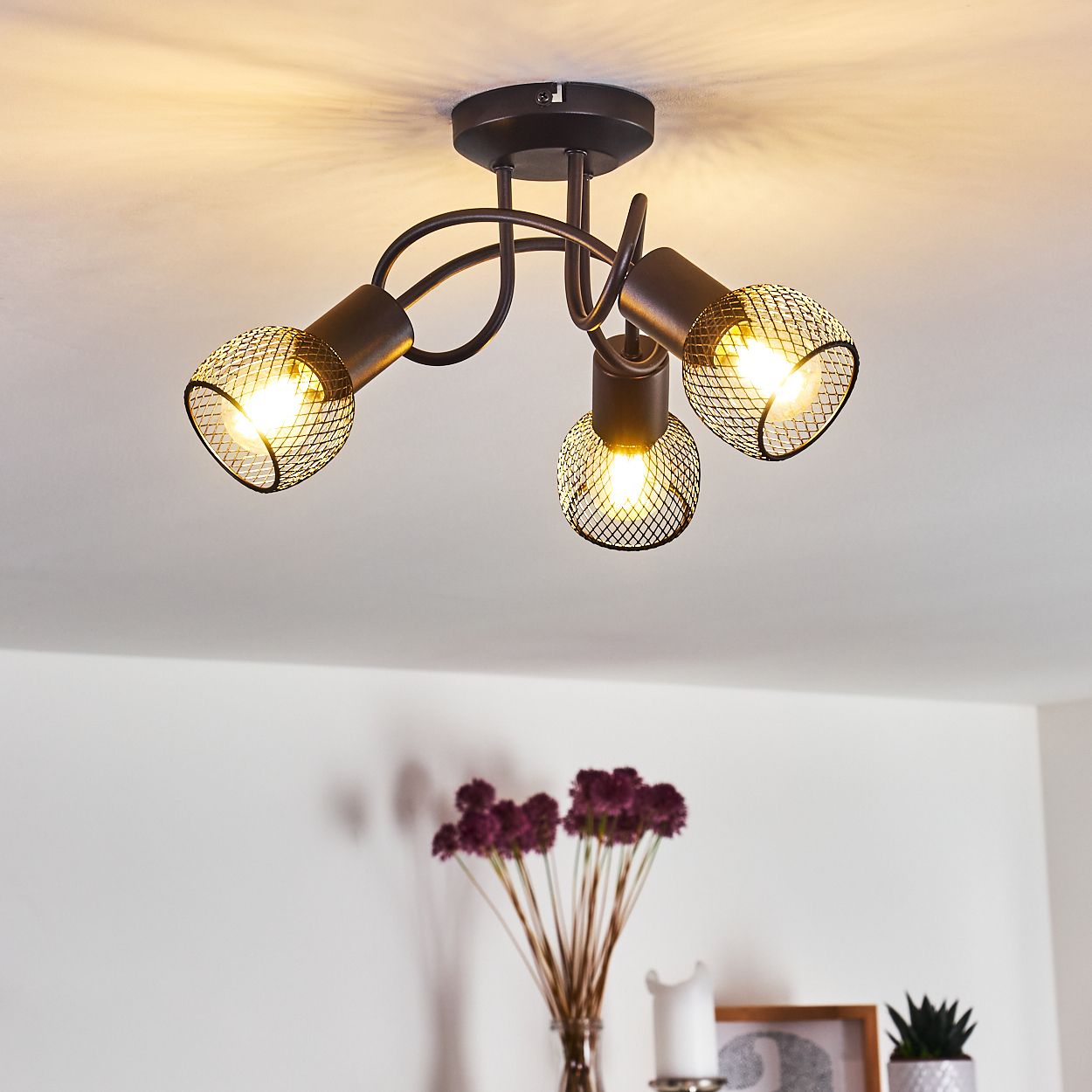 Eine Deckenleuchte mit drei lampen mit Gitterabdeckung. Darunter eine Vase mit lila Blumen und andere Dekorationselemente.
