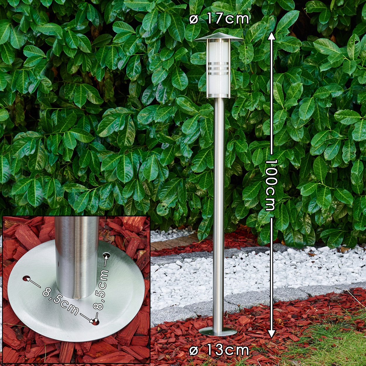 Eine Außenlampe aus Edelstahl. Sie steht hoch in einem Garten mit grünem Heckenhintergrund, roter Mulch und weißen Steinen.