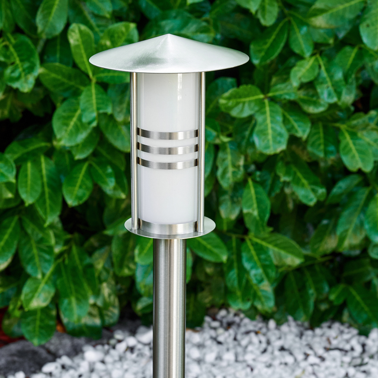 Eine moderne silberne Gartenlampe mit einem weißen Lichtzylinder. Grüne Blätter bilden den Hintergrund, weiße Kieselsteine sind zu sehen.