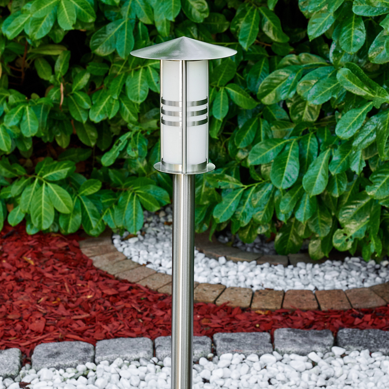 Eine moderne Gartenleuchte mit silberner Stange und Kappe. Grünes Laub im Hintergrund, roter Mulch, weiße Steine und ein Ziegelrand.