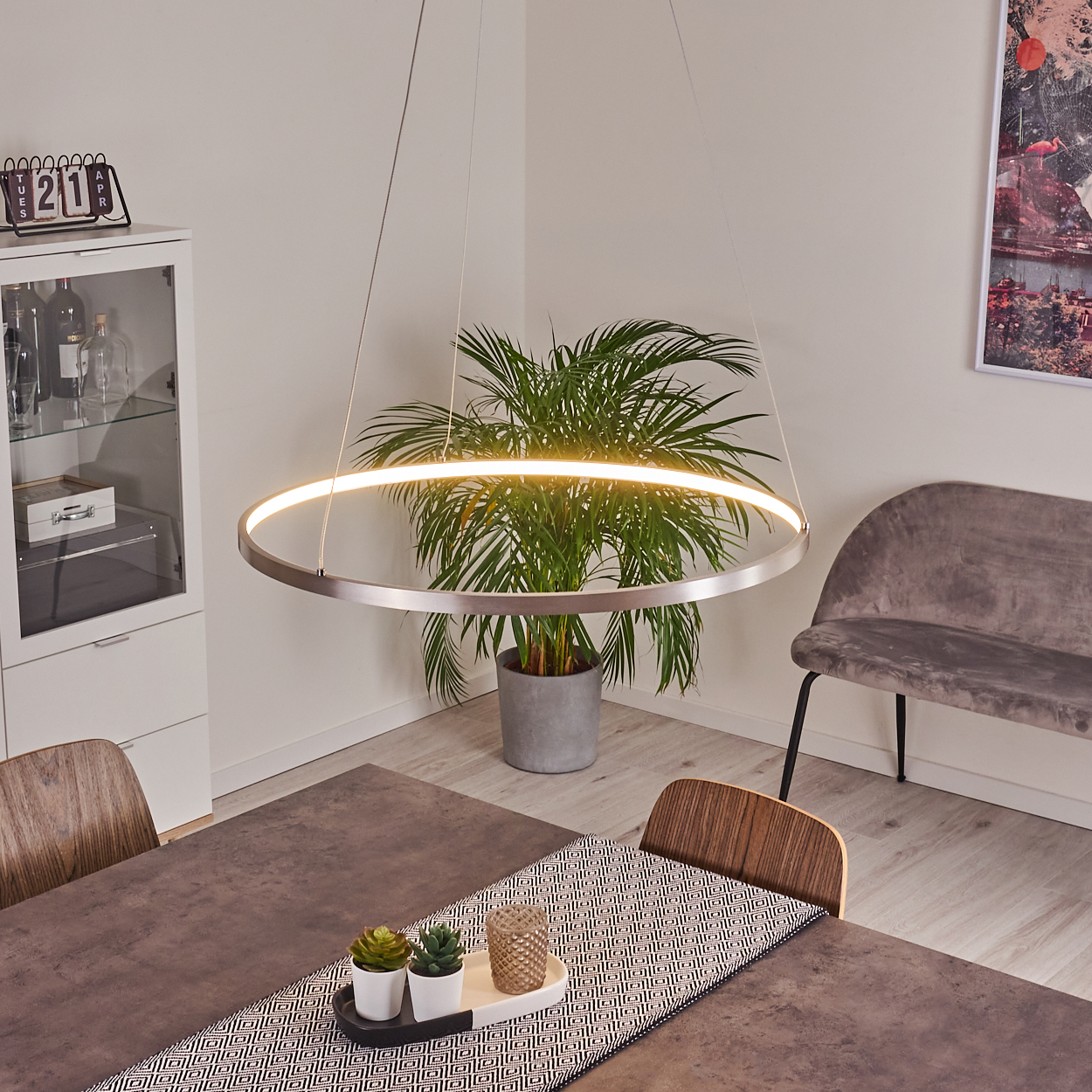 Ein modernes Esszimmer mit einer kreisförmigen Leuchte, einem Tisch mit Läufer, einer Pflanze und einem Samtstuhl.