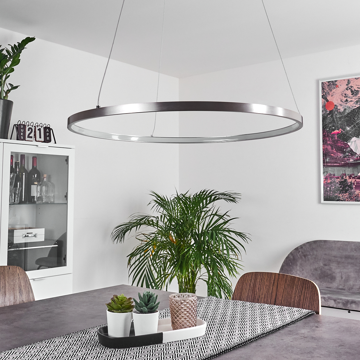 Ein Esszimmer mit einer großen kreisförmigen Leuchte. Ein Tisch ist mit Pflanzen und einem gemusterten Läufer gedeckt. Ein Schrank und Kunstwerke sind sichtbar.