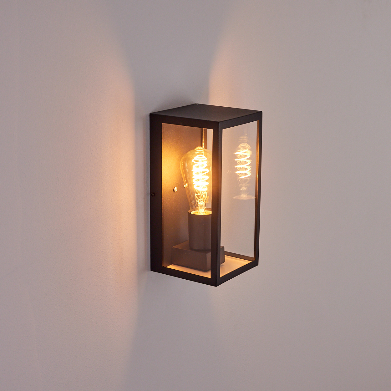 Eine schwarze, rechteckige Außenwandleuchte mit einer leuchtenden Edison-Glühbirne. Die Lampe ist an einer hellen Wand befestigt.