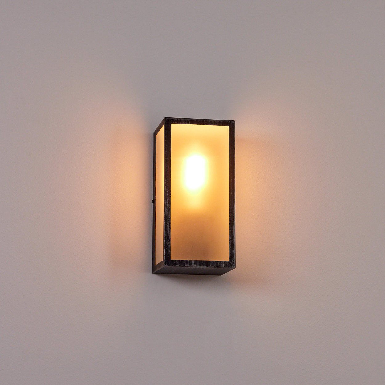 Eine rechteckige Wandleuchte mit dunklem Rahmen und Milchglas, die ein warmes, helles Licht an einer hellen Wand ausstrahlt.