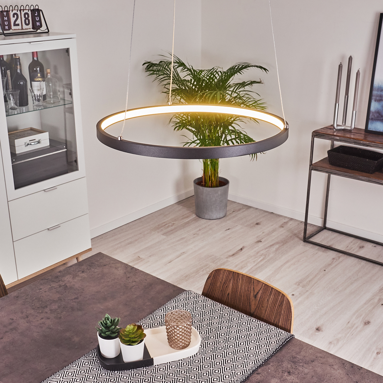 Ein modernes Esszimmer mit einer kreisförmigen Leuchte, einem Tisch mit gemustertem Läufer und einer Topfpflanze.