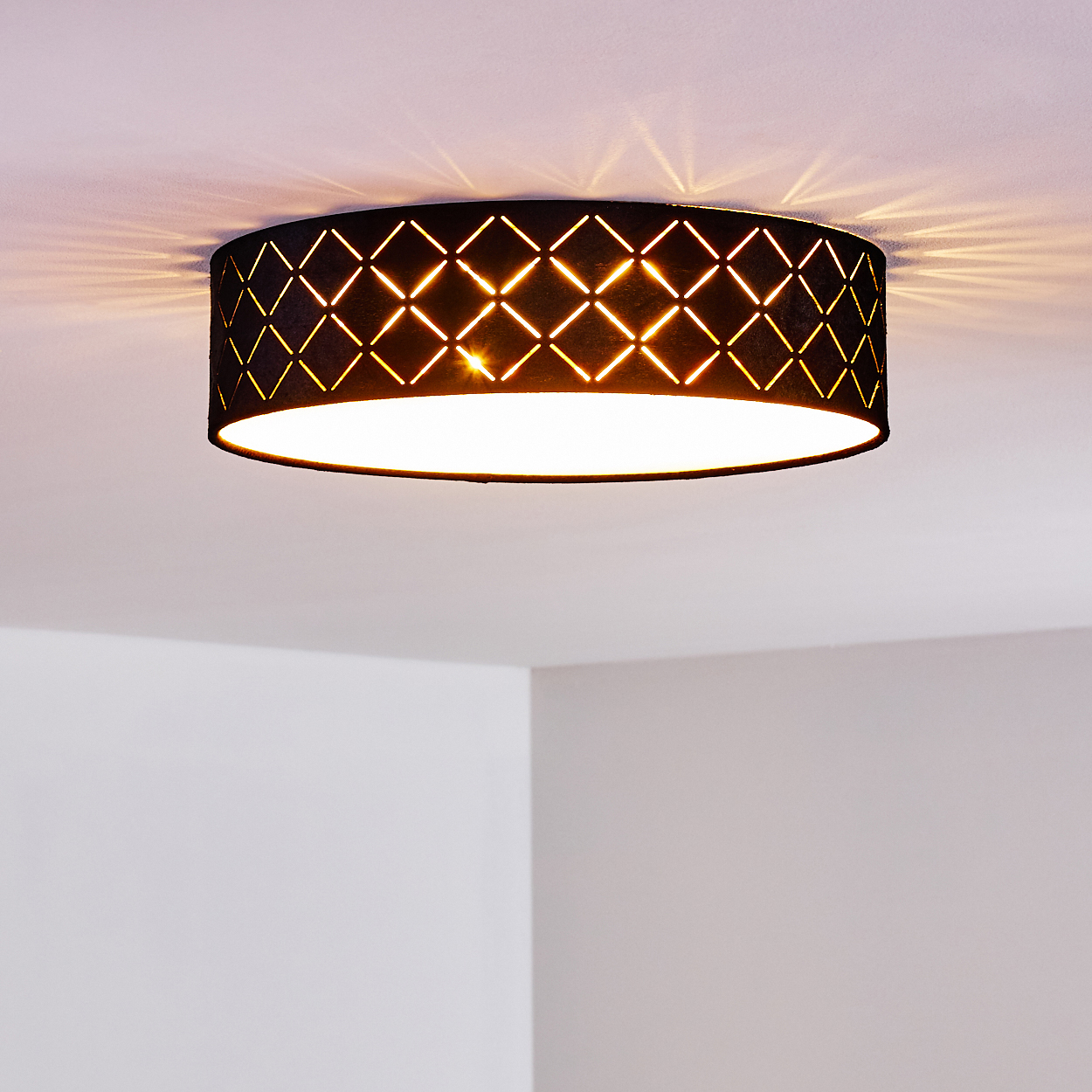 Eine schwarze Deckenlampe mit geometrischem Muster, die einen weißen Raum beleuchtet. Licht reflektiert sich an Decke und Wänden.