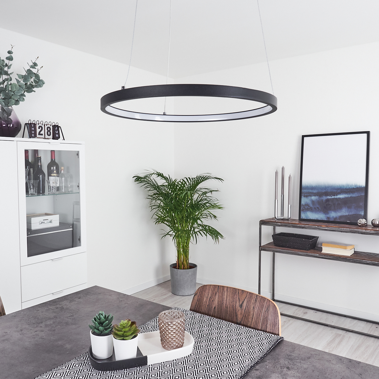 Modernes Esszimmer mit schwarzer kreisförmiger Leuchte, Pflanzen und einem Tisch mit Dekoration. Enthält einen Schrank und Kunstwerke.
