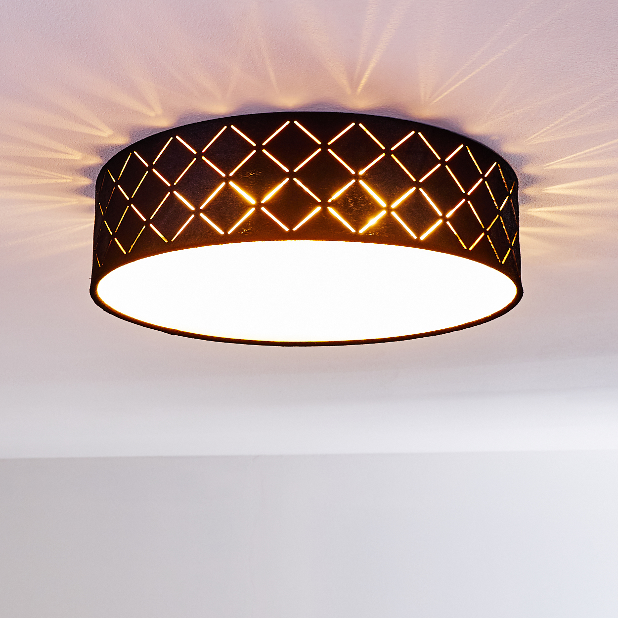 Eine schwarze Deckenlampe mit Diamantausschnitten. Licht scheint von der Lampe auf die Decke und beleuchtet den weißen Raum.