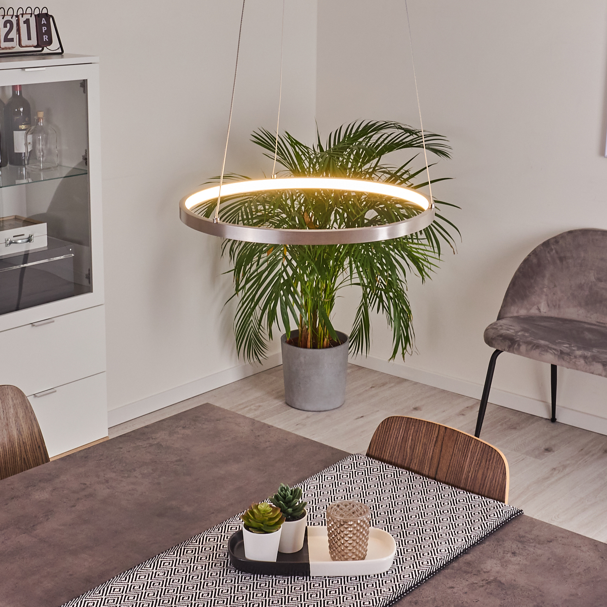 Ein modernes Esszimmer mit Tisch, Pflanzen, ringförmiger Lampe und Schrank. Ein Stuhl steht daneben, Tischdeko ist sichtbar.