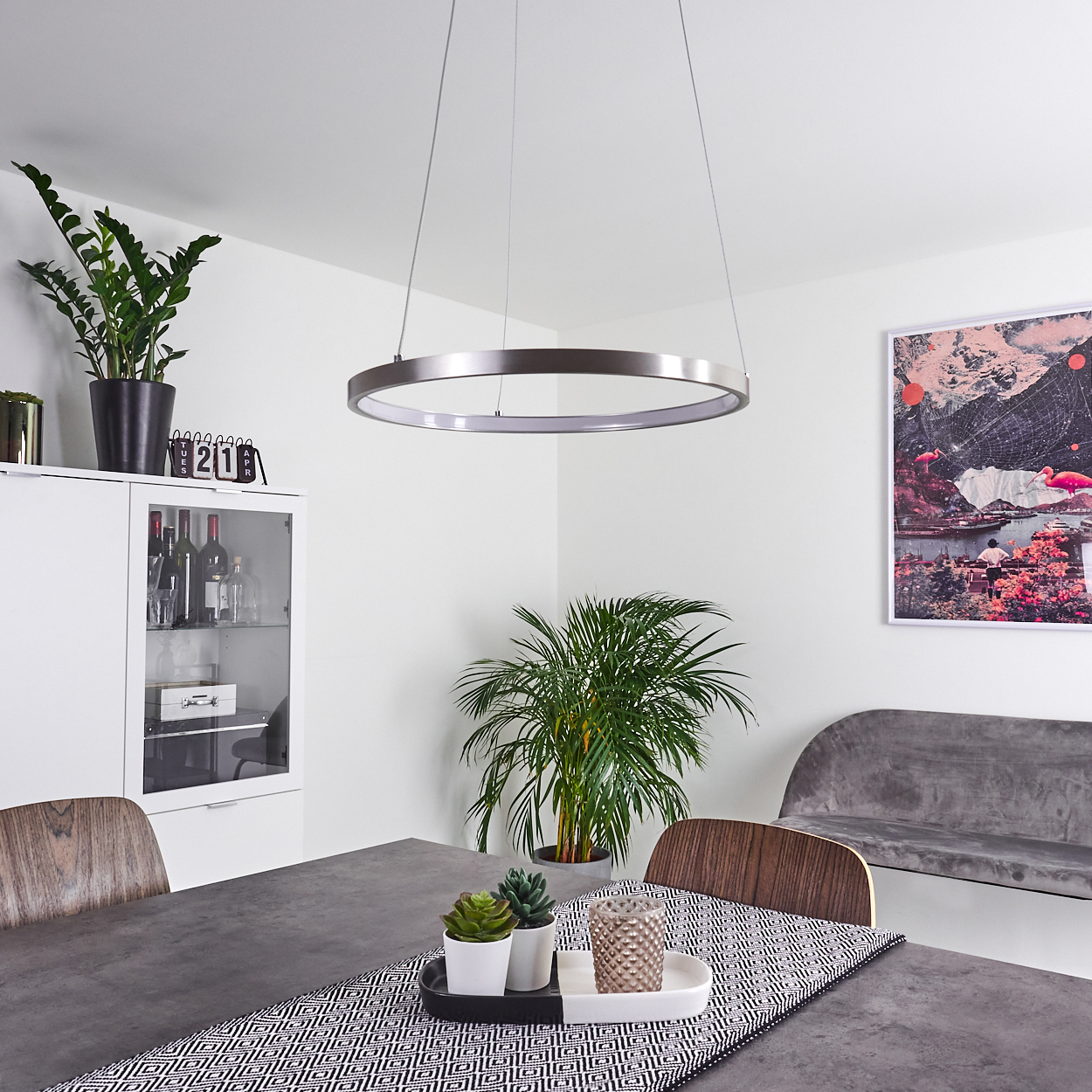 Ein Esszimmer mit modernem Design. Über einem dunkelgrauen Tisch hängt eine kreisförmige Leuchte. Pflanzen, Kunst und eine graue Bank sind sichtbar.