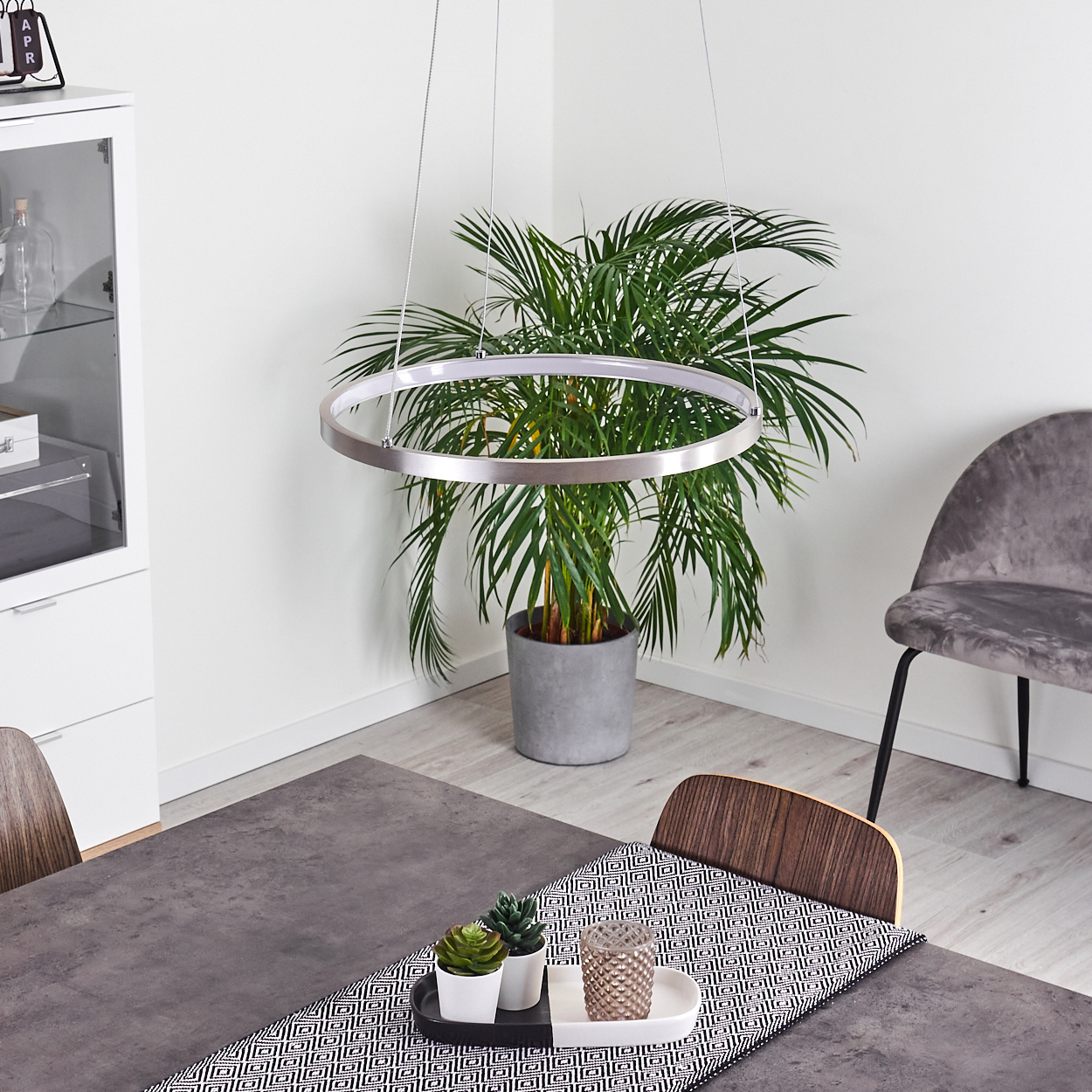 Ein modernes Esszimmer. Ein kreisförmiges Licht hängt über einem dunkelgrauen Tisch, der mit Pflanzen und einem gemusterten Läufer gedeckt ist. Im Hintergrund befindet sich eine große Pflanze.