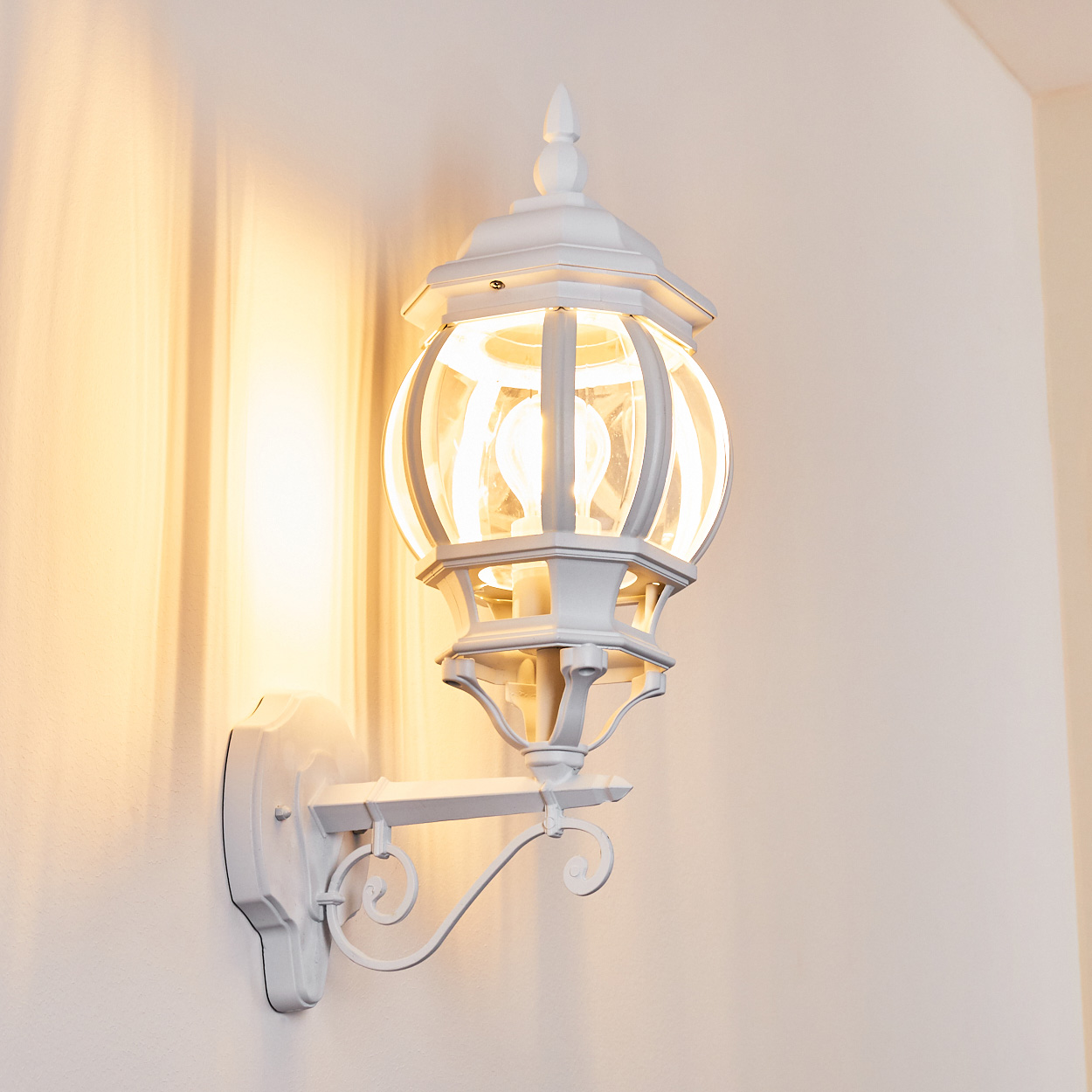Eine weiße Außenlampe ist an einer Wand befestigt. Sie hat ein rundes Glasgehäuse mit einer leuchtenden Glühbirne. Die Wand ist cremefarben.