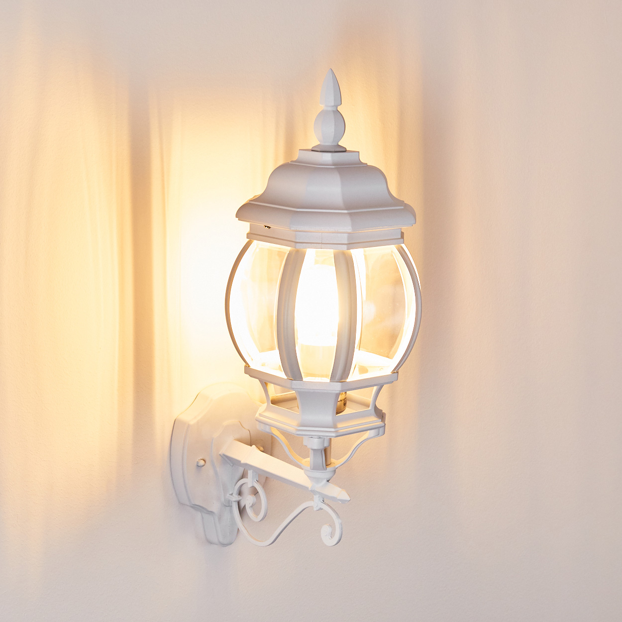 Eine weiße Außenlampe ist an einer Wand befestigt. Sie ist beleuchtet und das Licht wirft Schatten auf die Wand. Die Lampe hat ein klassisches Design.