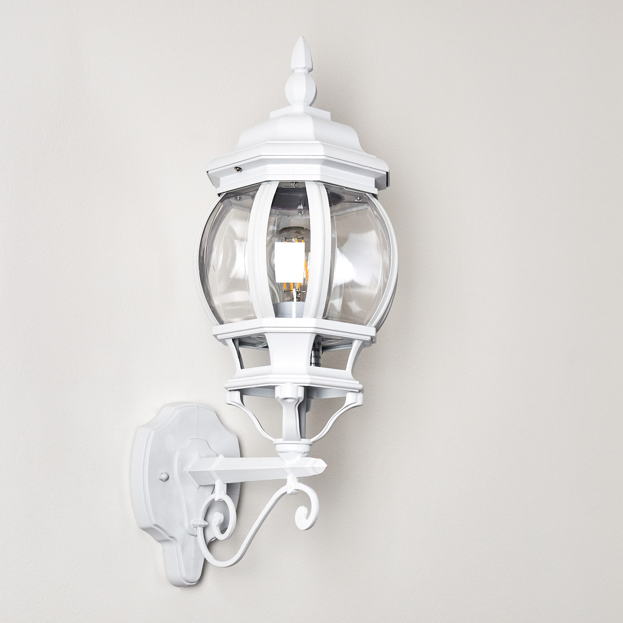 Eine weiße Außenlampe mit klarem Glaskugel und dekorativer Halterung ist an einer hellen Wand befestigt. Die Glühbirne leuchtet.