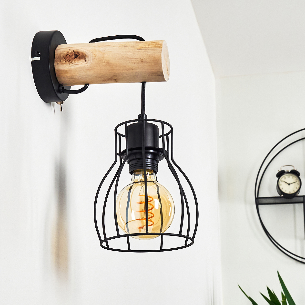 Eine Wandlampe mit einem schwarzen Käfig um eine Glühbirne, befestigt an einem Holzbalken und einer schwarzen Wandhalterung. Eine Uhr ist im Hintergrund.