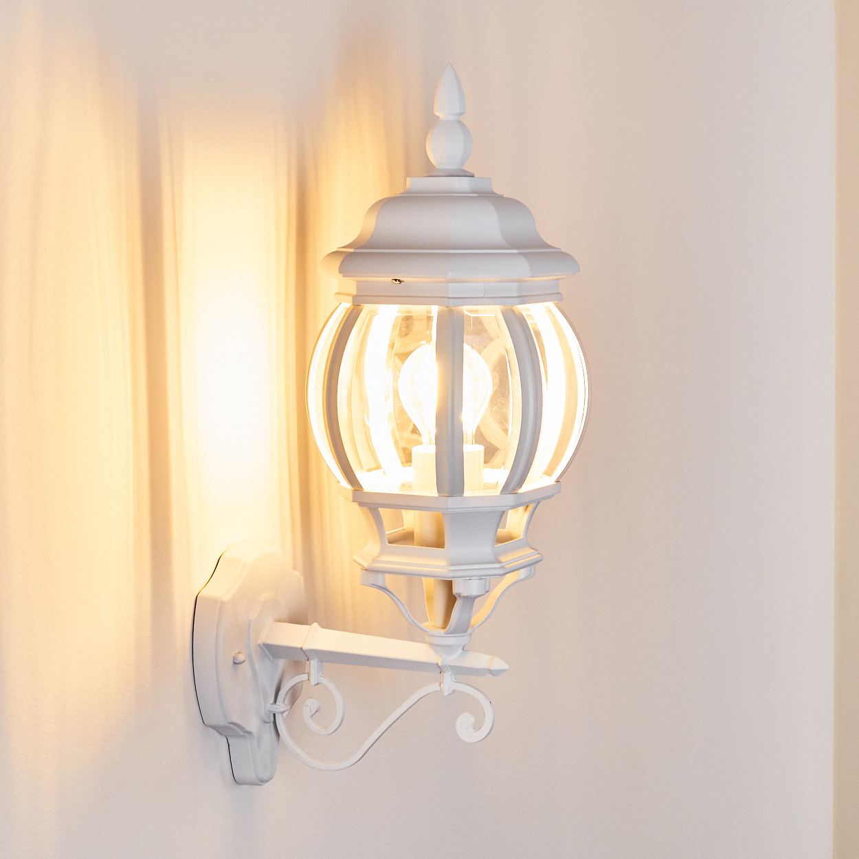 Eine weiße Außenwandleuchte mit Glaskugel und leuchtender Glühbirne, die ein warmes Licht auf die Wand wirft.