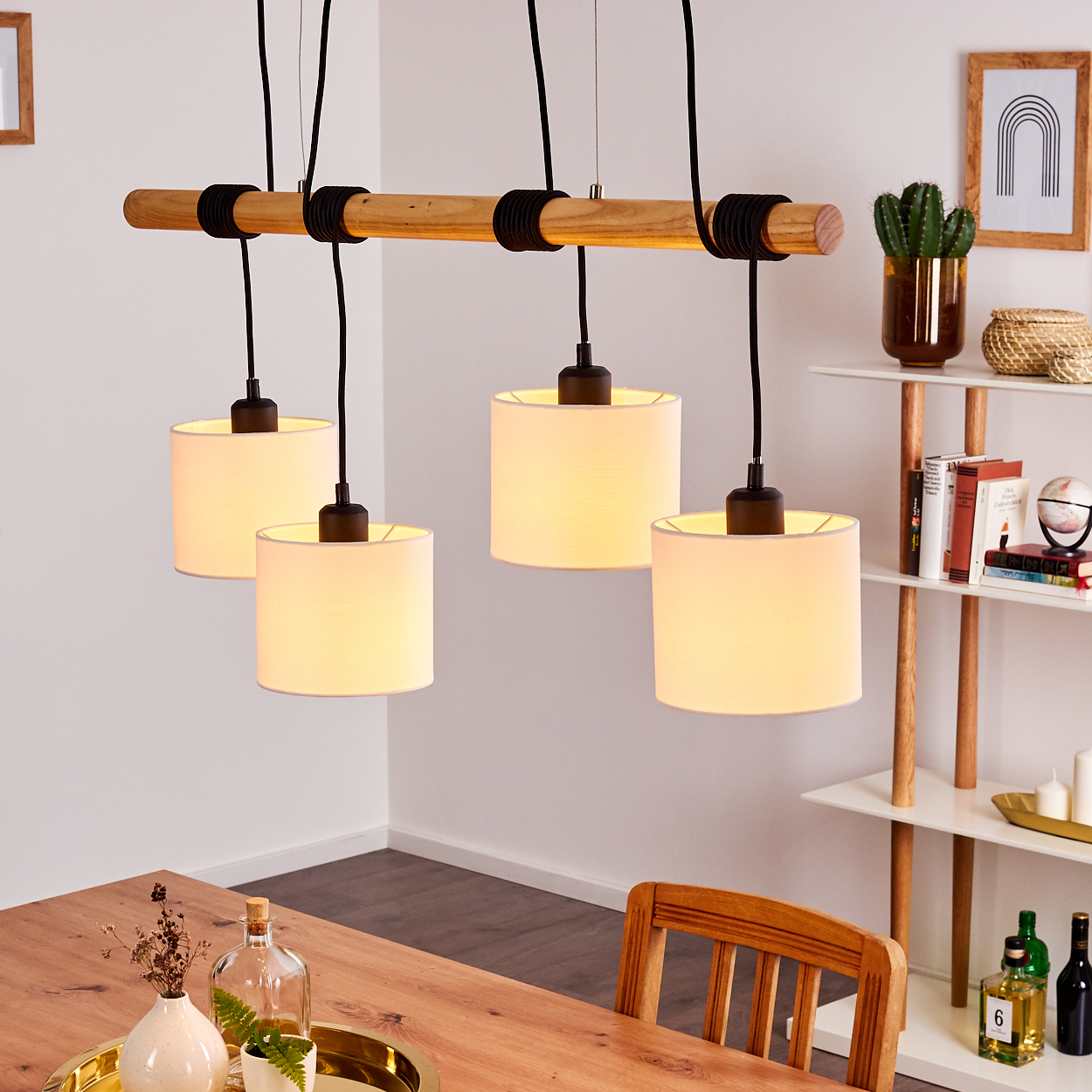 Vier Pendelleuchten hängen an einem Holzbalken. Ein Holztisch und ein weißes Regal mit Deko befinden sich im Raum.
