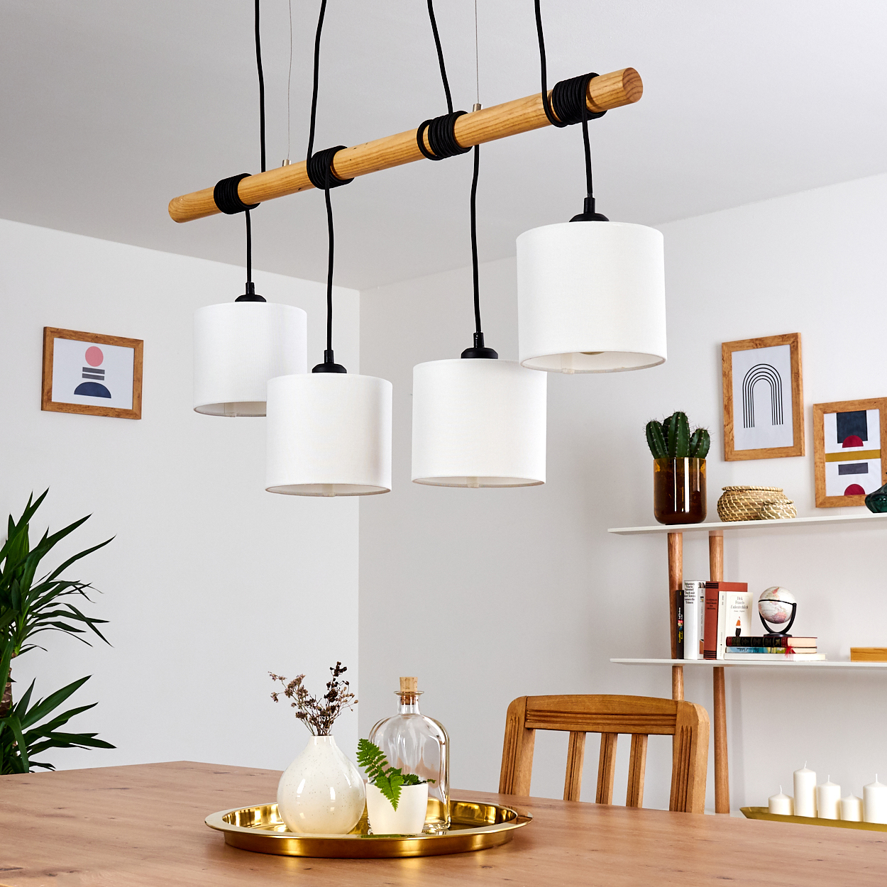 Ein Esszimmer mit Holztisch, weißen Lampen und Dekoartikeln. Ein Regal hält Bücher und Pflanzen. Weiße Wände und ein Holzstuhl.