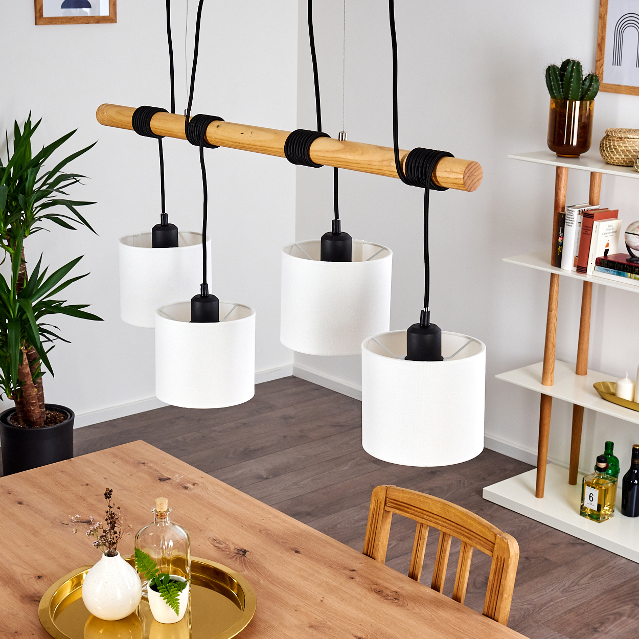 Ein Holzbalken mit vier weißen Lampen hängt über einem Holztisch. Im Hintergrund befinden sich eine grüne Pflanze und ein weißes Regal mit Dekoration.