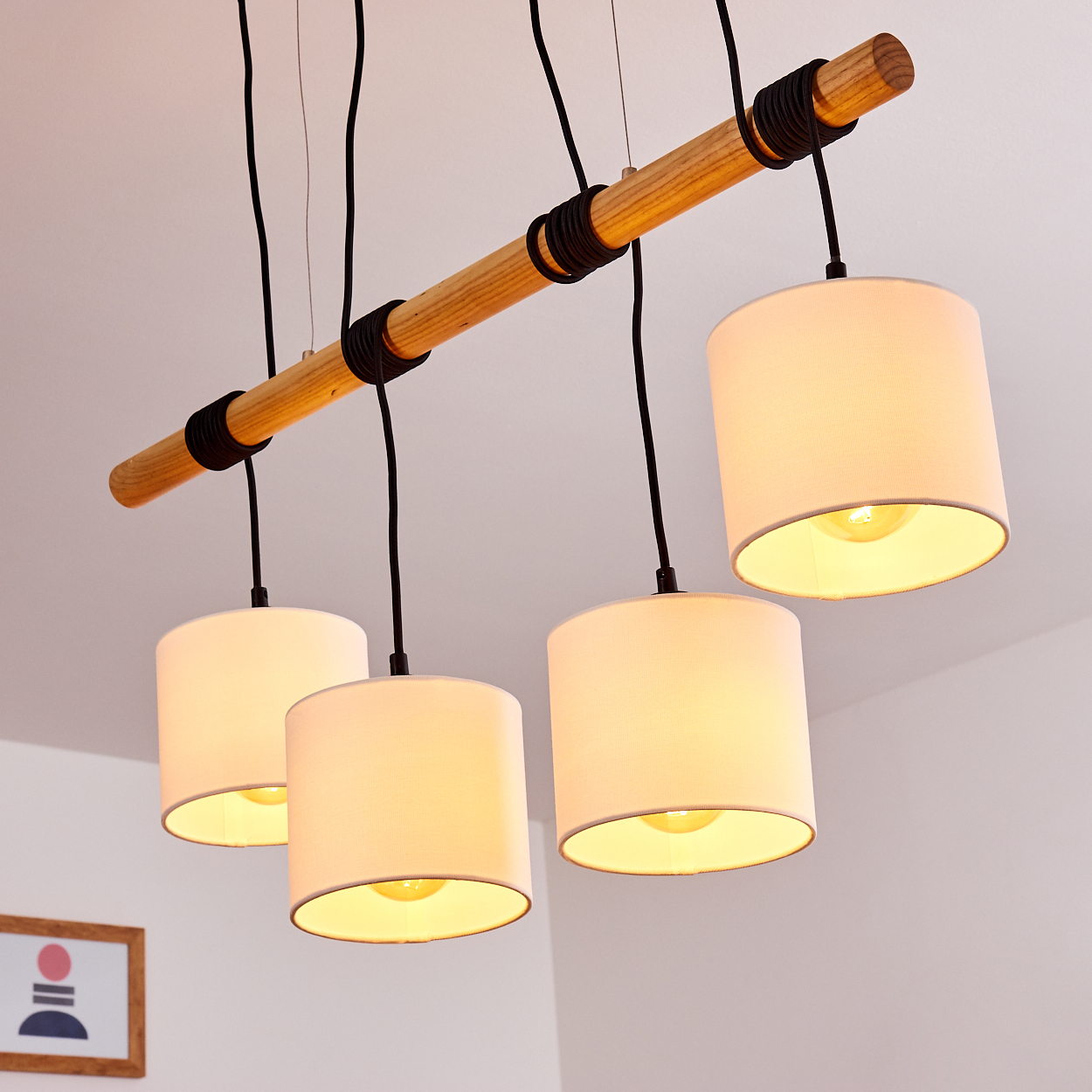 Vier Pendelleuchten hängen von einem Holzbalken. Die Schirme sind weiße Zylinder, die warmes Licht aussenden. Schwarze Kabel und Akzente sind sichtbar.