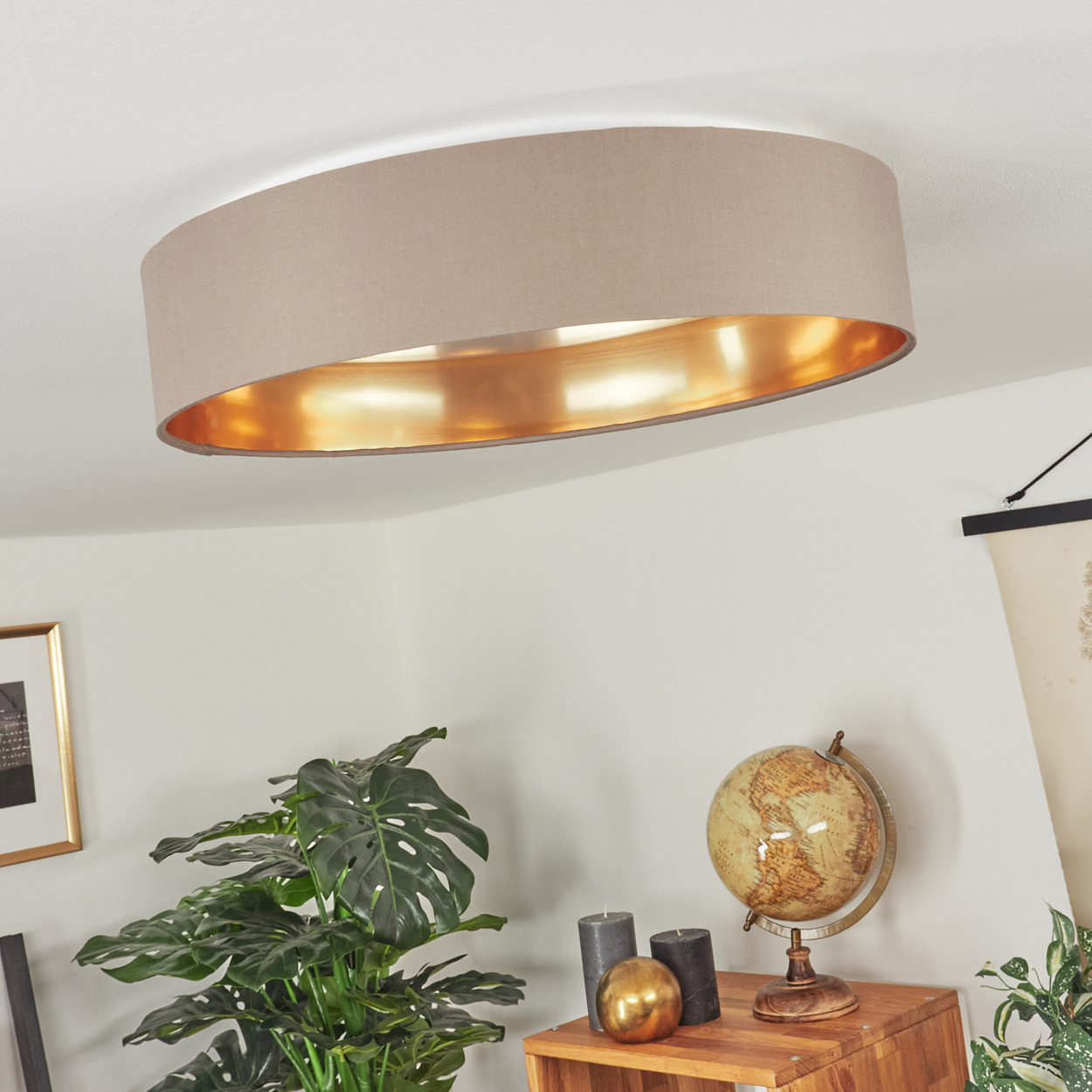 Eine große, beige, ovale Deckenleuchte mit goldenem Innenraum. Darunter ein Holztisch mit Globus, Kerzen und Pflanzen.
