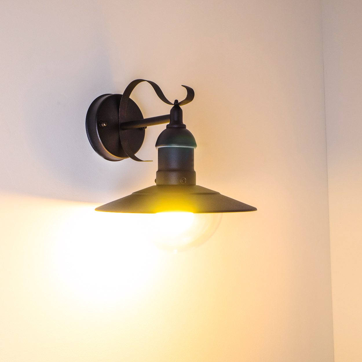 Eine schwarze Wandleuchte mit nach unten gerichteten Schirm wird beleuchtet. Der Hintergrund ist eine weiße Wand.