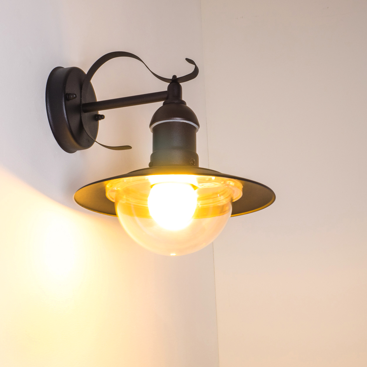 Eine schwarze Wandlampe mit Glasschirm ist an einer weißen Wand montiert. Das Licht ist an und wirft einen warmen Schein.