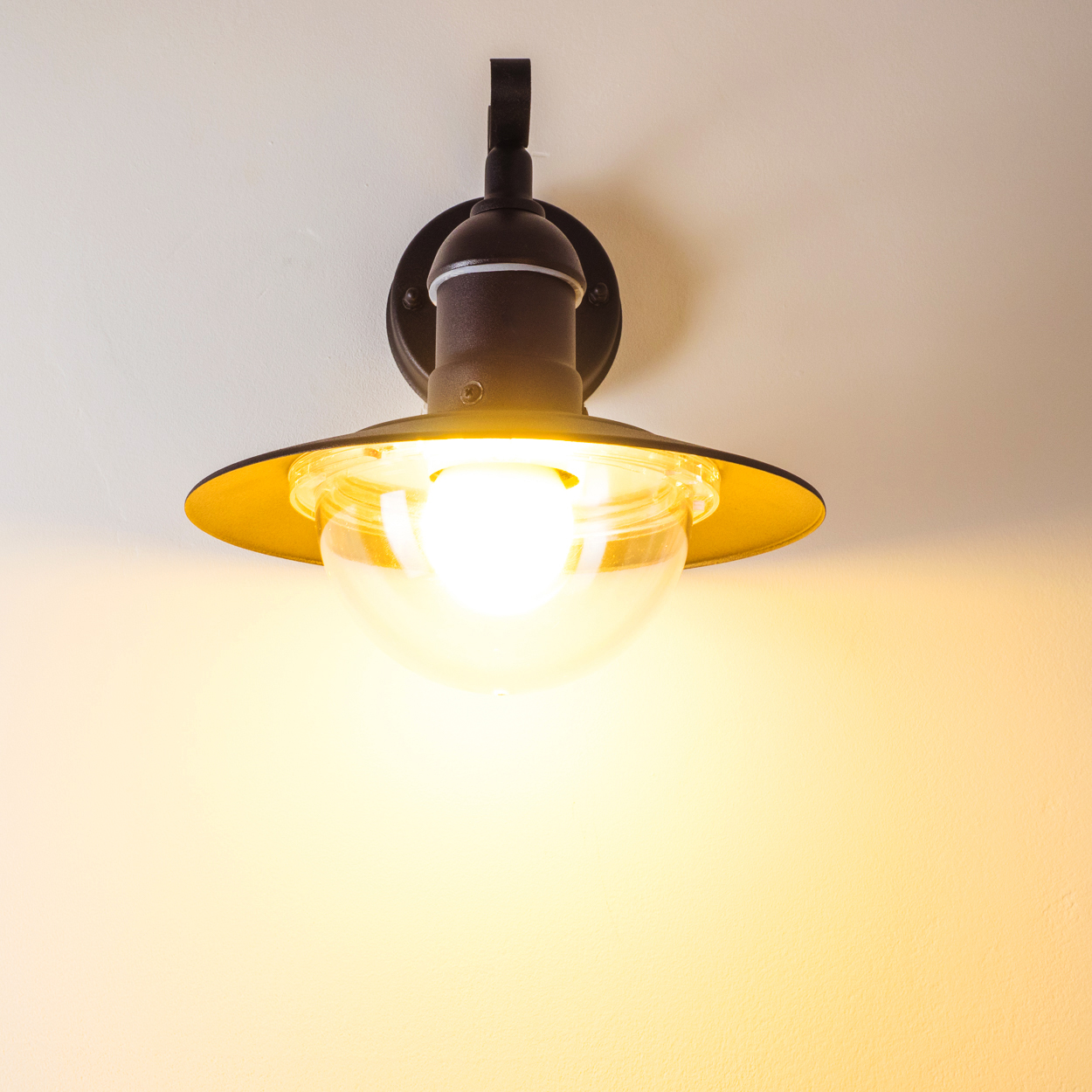 Eine Wandleuchte mit runder Glasabdeckung und goldfarbenem Schirm. Die Lampe befindet sich an einer weißen Wand und leuchtet.