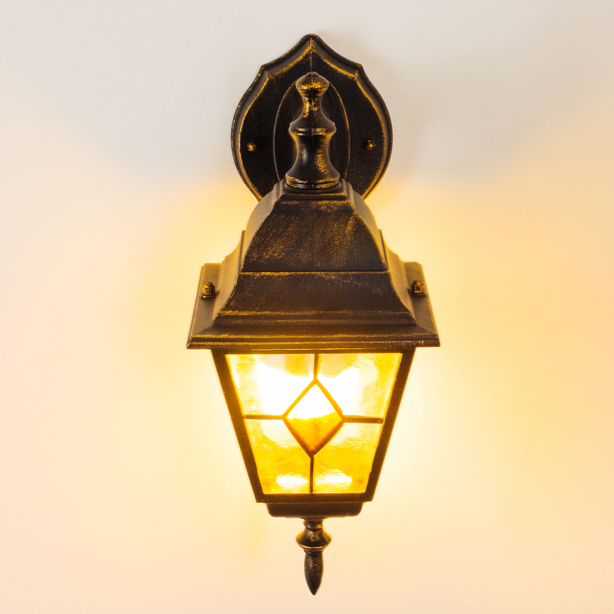 Eine verzierte, schwarz-goldene Wandlampe mit gelbem Licht. Sie ist an einer cremefarbenen Wand befestigt.