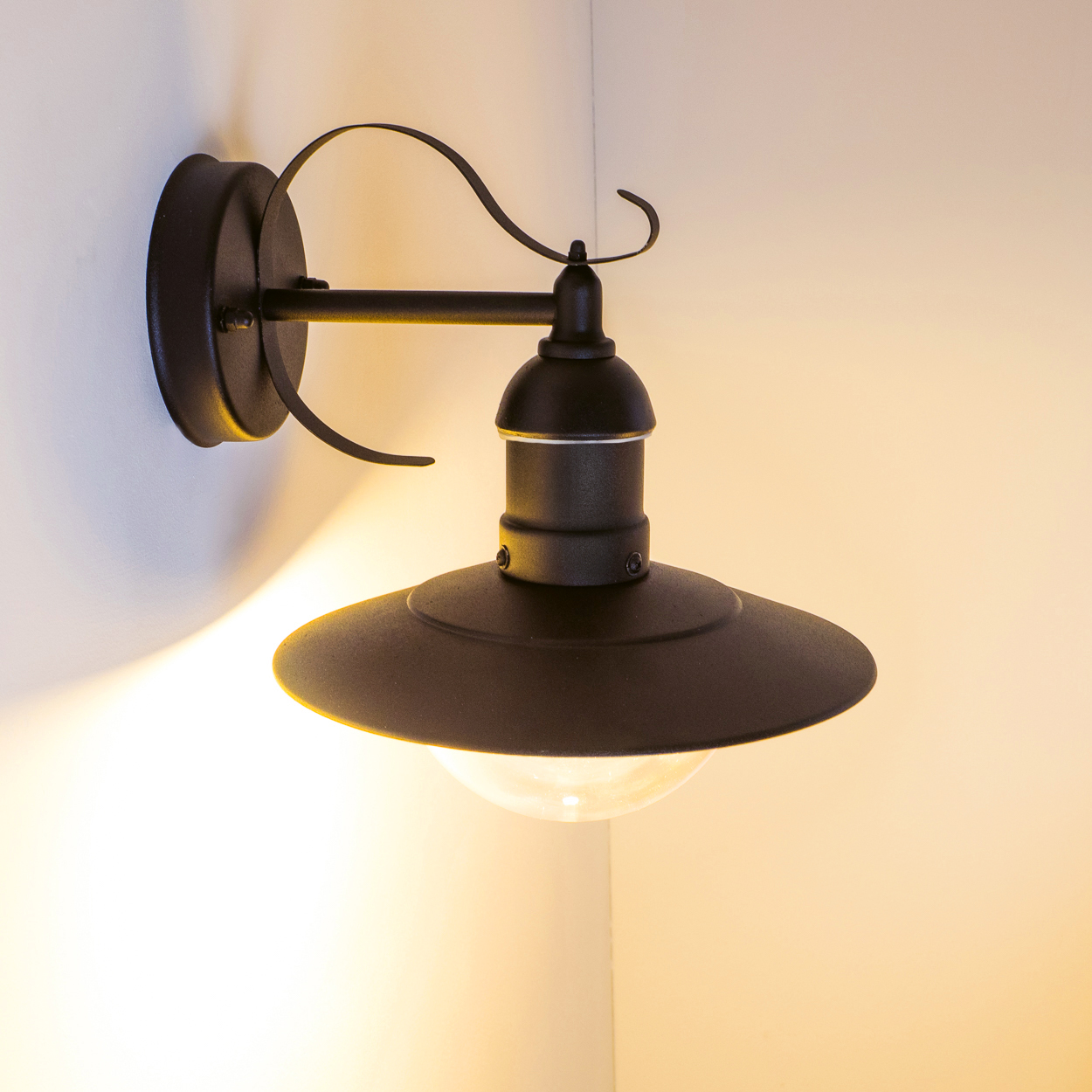 Eine schwarze Wandlampe mit rundem Schirm und klarer Glühbirne ist an einer weißen Wand montiert und wirft einen warmen Schein.