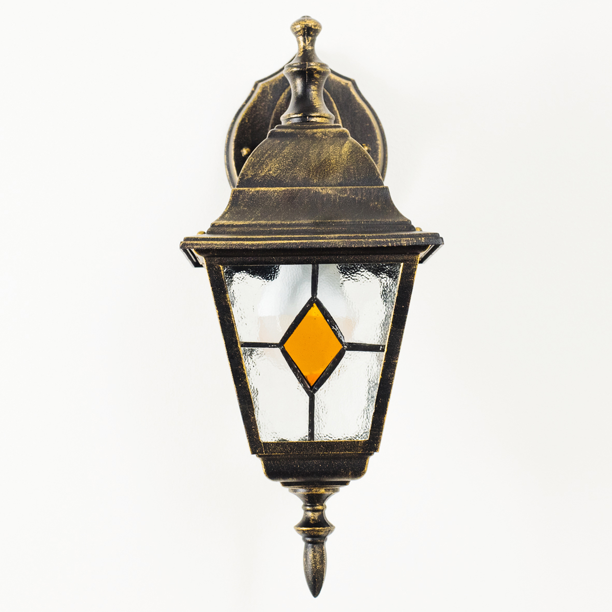Eine verzierte Außenlampe mit einem rautenförmigen Glas in Bernstein, vor weißem Hintergrund. Die Lampe ist bronzefarben.