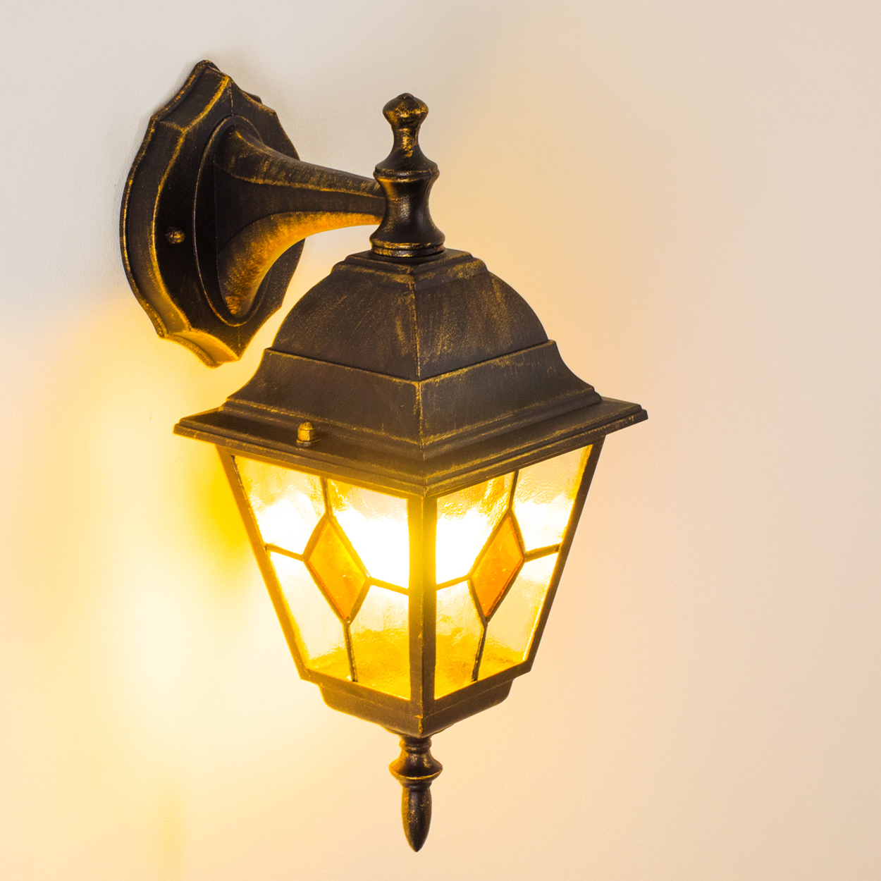 Eine verzierte, goldene Außenlampe an der Wand. Die Lampe ist beleuchtet, mit rautenförmigen Glasscheiben.