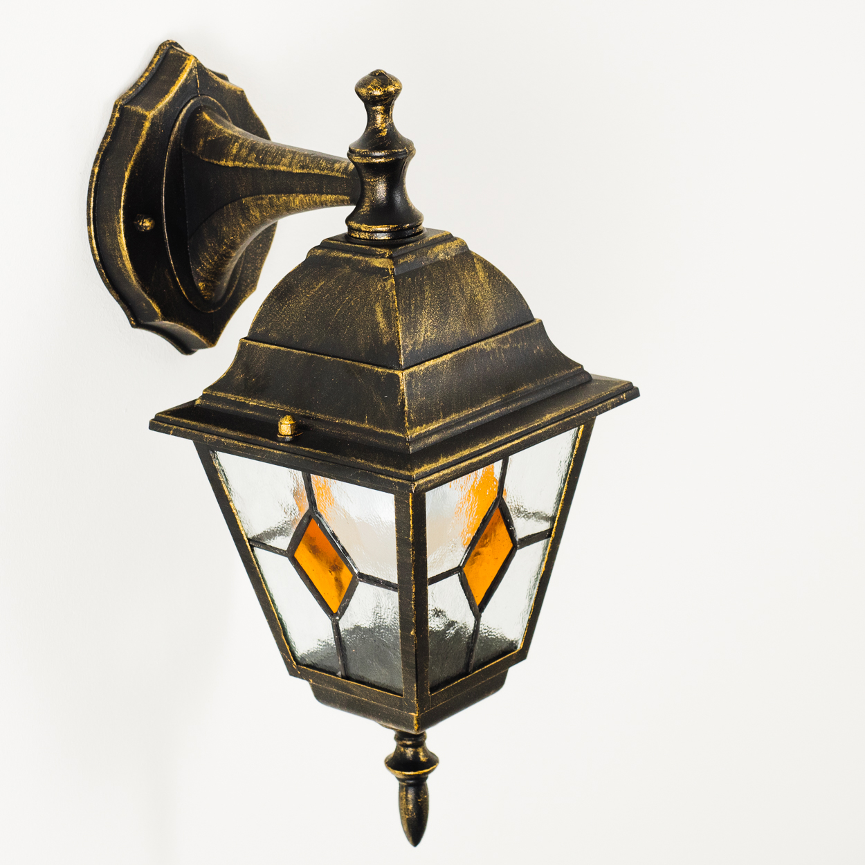 Eine verzierte Wandlampe im antiken Stil. Sie hat eine schwarz-goldene Oberfläche, Glasscheiben und eine dekorative Spitze. Sie ist an einer weißen Wand befestigt.